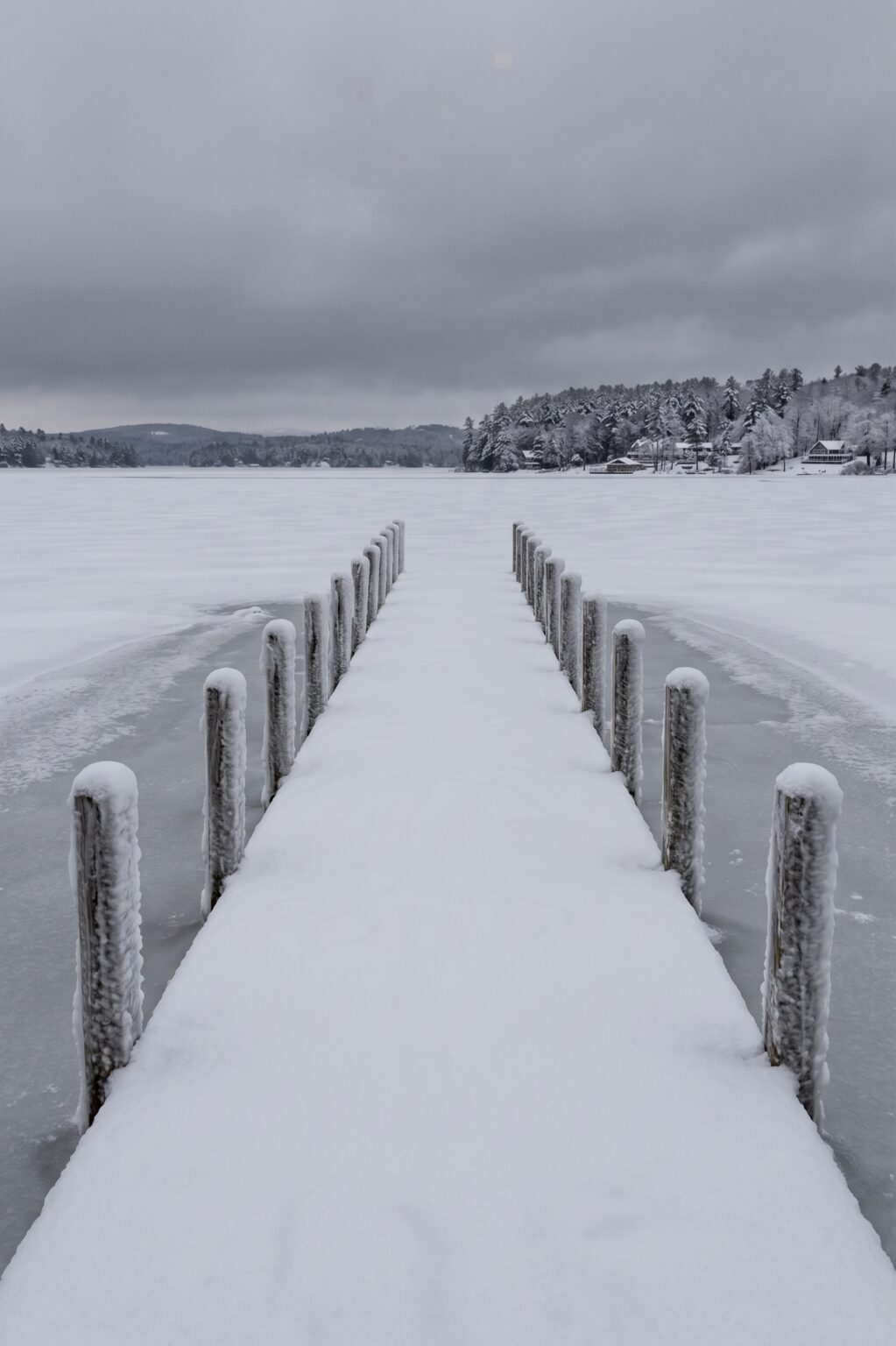 A snow-covered dock lined with ice-crusted pilings running out onto a frozen lake under heavy grey cloud.